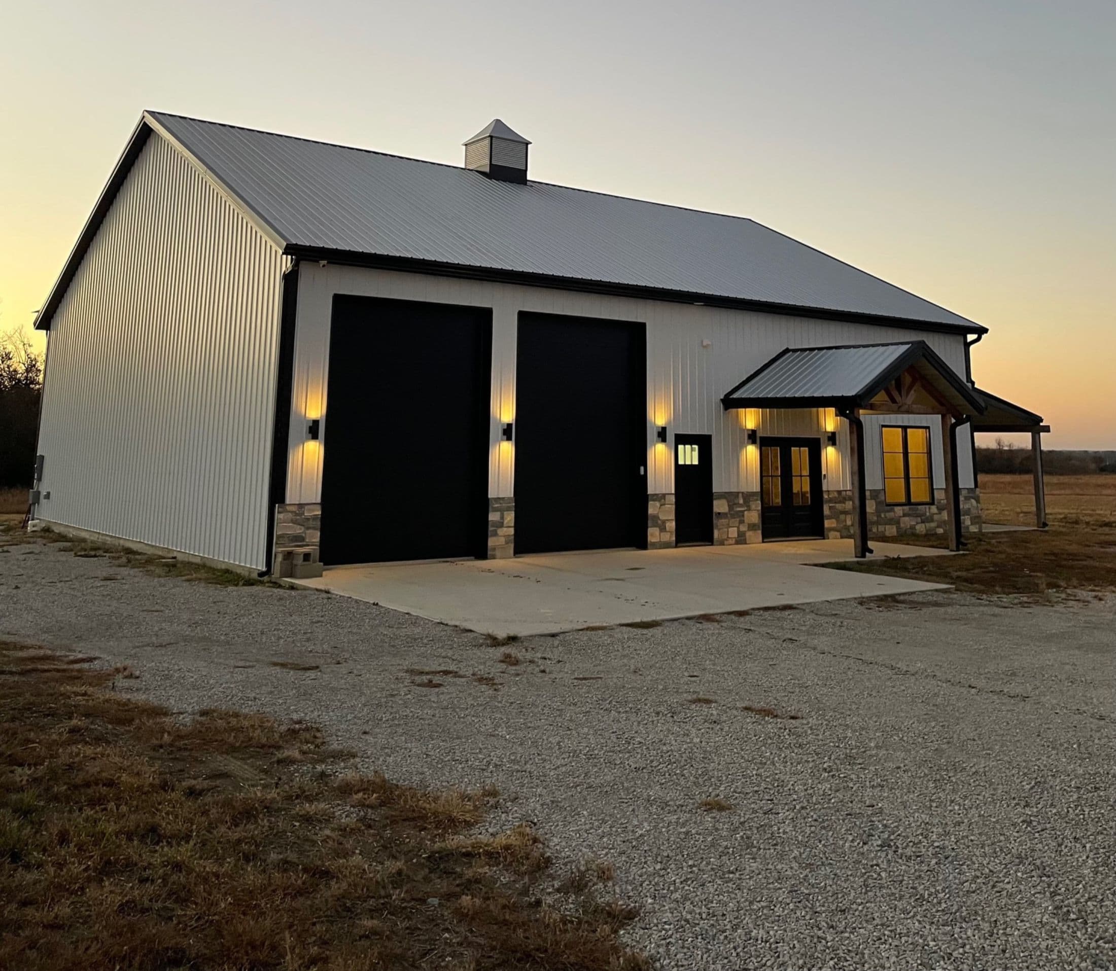 White barndominium with black overhead doors and stone accents at dusk